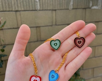 May include: Four gold necklaces with heart-shaped pendants. Each pendant features a concentric circle design in different colors: red, blue, green, and pink.