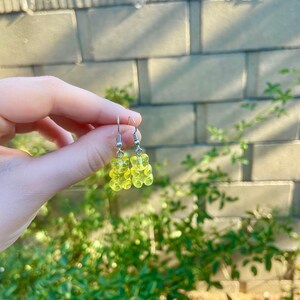 K&ouml;nnte beinhalten: Ein Paar gelbe Gummib&auml;rchen-Ohrringe mit silbernen Haken. Die Ohrringe h&auml;ngen an der Hand einer Person.