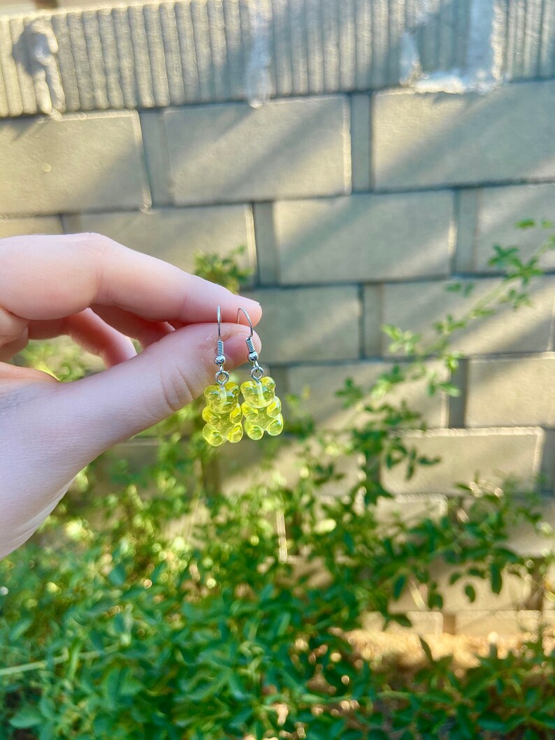 K&ouml;nnte beinhalten: Ein Paar gelbe Gummib&auml;rchen-Ohrringe mit silbernen Haken. Die Ohrringe h&auml;ngen an der Hand einer Person.