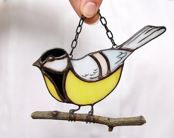 May include: Stained glass bird ornament in yellow, white, black, and pale pink. The bird is perched on a small wooden branch and suspended by a black chain. The bird's design features a yellow breast, white wings, and a black head.