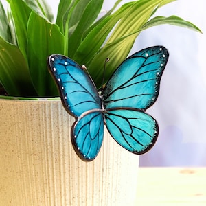 Peut inclure: Un pot en céramique blanc avec une décoration de papillon bleu. Le pot a une surface texturée et trois pieds. Une plante verte pousse dans le pot.