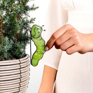 May include: A green metal garden stake shaped like a smiling caterpillar with black eyes and a red mouth. The stake is being held by a person's hand.
