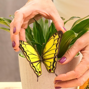 May include: A yellow butterfly decoration with black outlines and red accents. The butterfly is held near a green plant in a beige pot. The butterfly's wings are a bright yellow color.