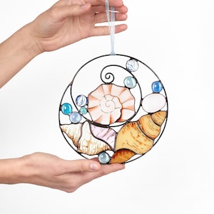 Peut inclure: Un attrape-soleil en vitrail avec un motif circulaire de coquillages et de perles de verre bleues. Les coquillages sont dans des tons de brun, rose et blanc.