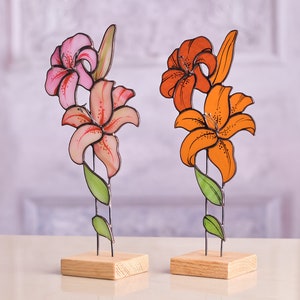 Op de afbeelding: Twee lelie-sculpturen van glas-in-lood op lichte houten voetstukken. Het ene beeld heeft roze en licht perzikkleurige bloemblaadjes met donkere spikkels, terwijl het andere levendige oranje bloemblaadjes met donkere spikkels heeft. Beide hebben groene glazen bladeren.