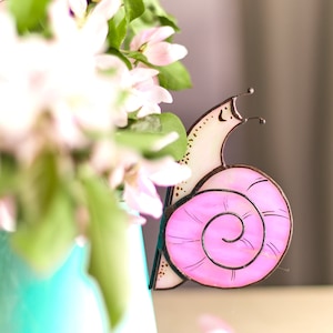 May include: A pink and white stained glass snail with a brown outline. The snail is sitting on a table with a bouquet of white flowers in the background.