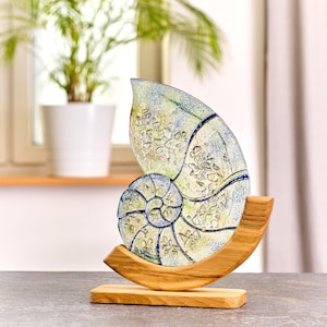 May include: A glass nautilus shell sculpture with blue and green swirls on a wooden base. The shell is translucent and has a detailed design.