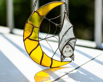 May include: A stained glass hanging decoration featuring a yellow crescent moon with a white bat hanging upside down from it. The bat has a web behind it and black eyes.