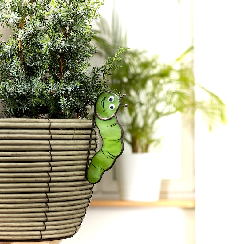 Op de afbeelding: Een groene keramische rups met oogjes zit op de rand van een geweven rieten pot. De pot is gevuld met een groene, bladerrijke plant.