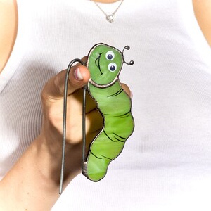Op de afbeelding: Een groene, van glas gemaakte rups met een glimlachend gezicht en knoopjesogen. De rups is bevestigd aan een metalen draad.