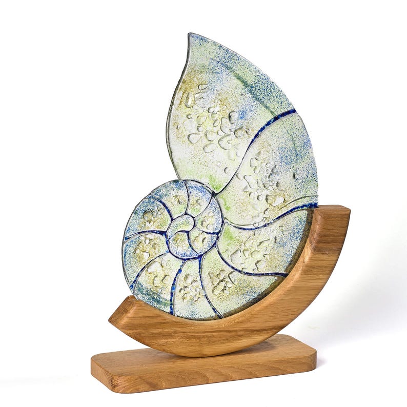 A handcrafted fused glass nautilus shell sculpture with textured details in soft greens and blues, displayed on a curved wooden base, perfect for coastal-inspired home décor.
