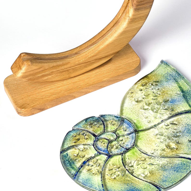 May include: A wooden stand with a curved top holds a glass nautilus shell with a blue and green mosaic design. The shell has a textured surface with small bubbles.