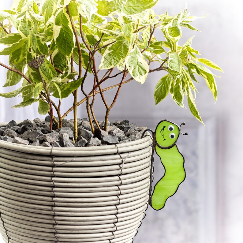 Op de afbeelding: Een witte rieten plantenbak met een groene glazen glimlachende rupsdecoratie. De plantenbak is gevuld met grijs grind en een groen-witte bonte plant.