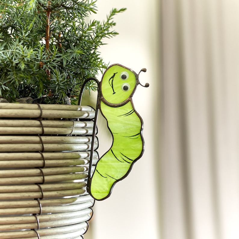 Op de afbeelding: Een groene glazen rups met goggle-ogen en een glimlachend gezicht is bevestigd aan een rieten plantenpot.