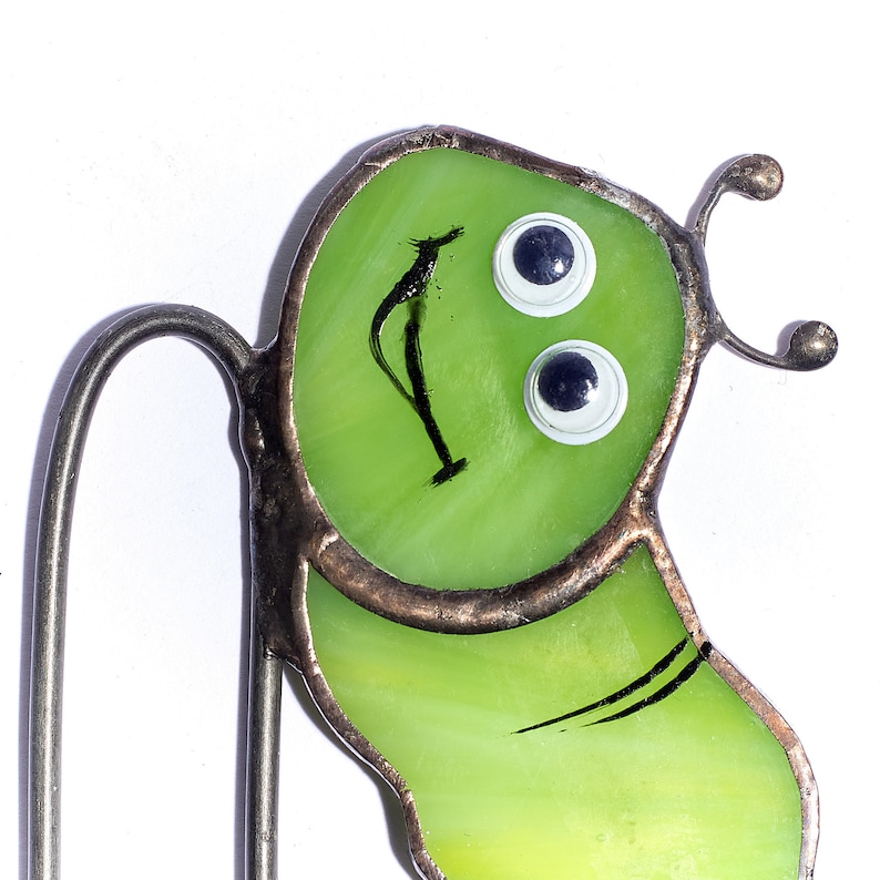 Op de afbeelding: Een groene glas-in-lood bladwijzer in de vorm van een glimlachende rups met zwarte ogen en een zwarte glimlach. De bladwijzer heeft een metalen draad die aan de bovenkant is bevestigd.