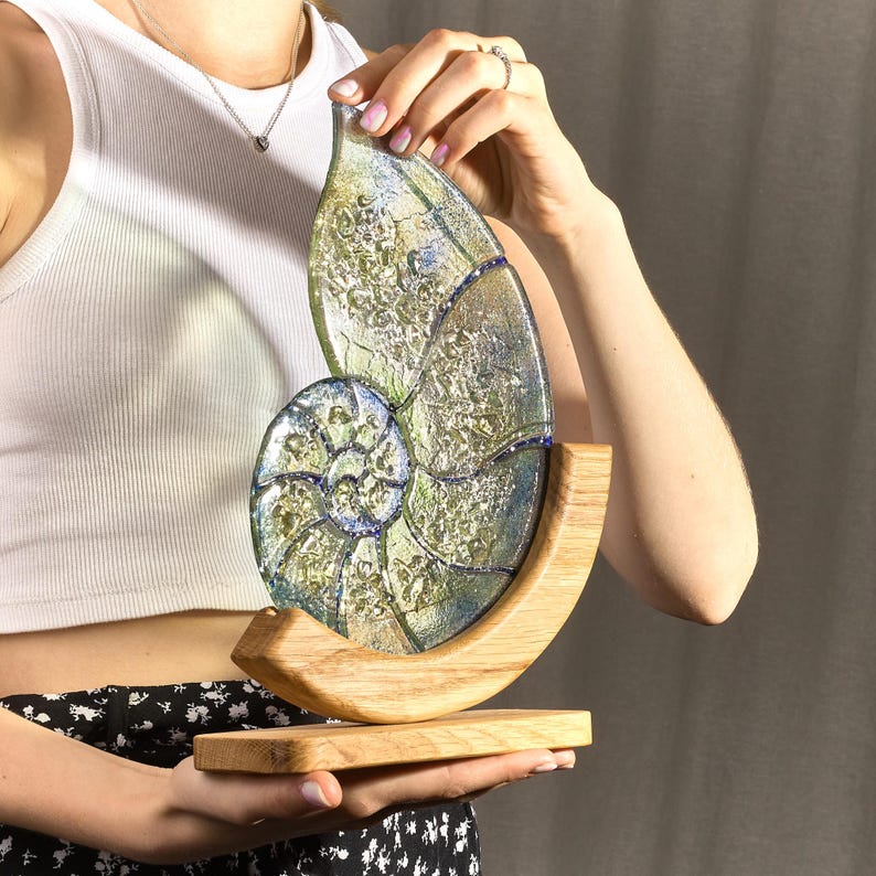 May include: A glass sculpture of a nautilus shell with iridescent green and blue colors. The shell is mounted on a wooden base.