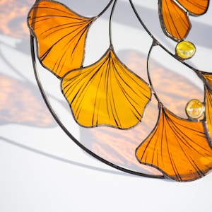 Op de afbeelding: Een glas-in-loodraam met een cirkelvormig ontwerp van meerdere ginkgo-bladeren in amber- en gele tinten. De bladeren zijn zwart omrand en hebben een delicaat adernetwerk.
