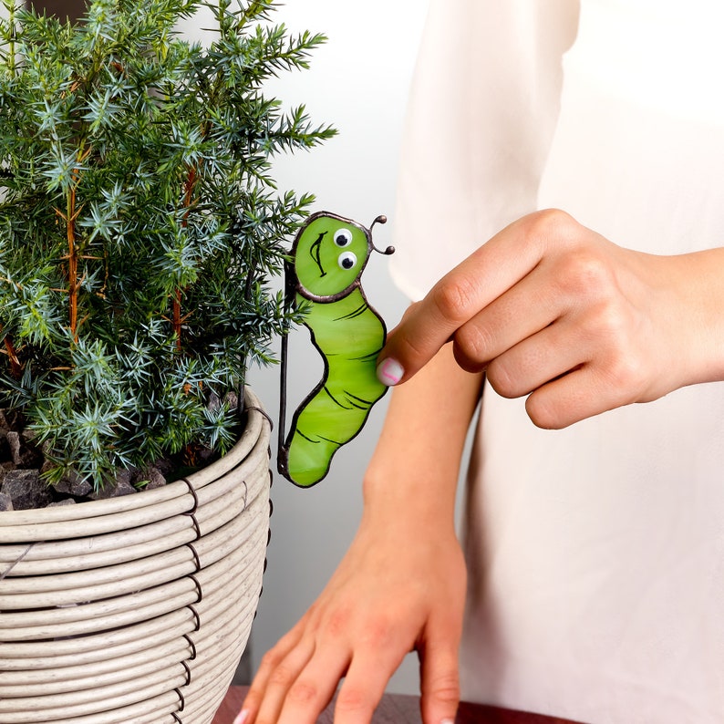 Op de afbeelding: Een groene glazen moza&iuml;ekrups met een glimlachend gezicht en zwarte ogen. De rups is bevestigd aan een metalen staaf en wordt vastgehouden door een hand.