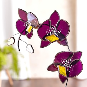 May include: A stained glass orchid with purple petals, yellow centers, and white accents. The orchid is mounted on a wire stem.