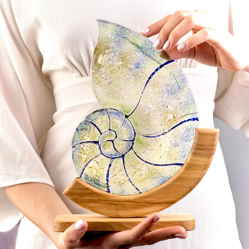 May include: A nautilus shell sculpture made of stained glass with blue, green, and yellow tones. The shell is mounted on a wooden base.