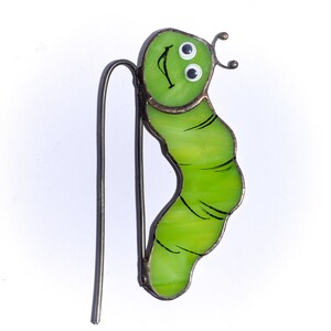 Op de afbeelding: Een groene glas-rups met zwarte ogen en een glimlachende mond. De rups is bevestigd aan een metalen draad.