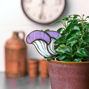 May include: A purple and white stained glass mushroom plant stake. The stake is inserted into a terracotta pot with a green plant.