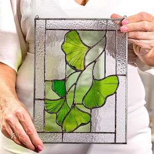 May include: A stained glass window panel featuring a design of green ginkgo leaves in a geometric pattern. The leaves are outlined in brown and set against a clear glass background.