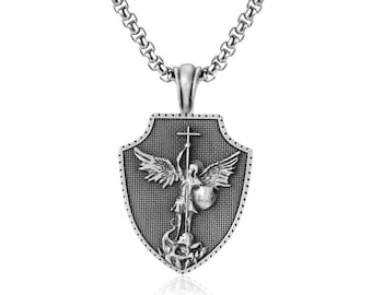 Può includere: Collana con pendente color argento con un pendente a forma di scudo con una figura di angelo, una croce e uno scudo. Il pendente è appeso a una catena con un design a maglie arrotondate e strutturate. L'angelo è raffigurato con ali e una spada.