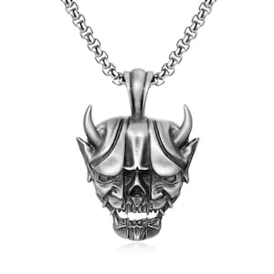May include: Silver-toned pendant necklace featuring a detailed oni mask design. The pendant has curved horns, a grimacing mouth, and intricate facial features. The chain is a linked design, and the overall style is gothic.