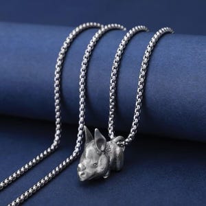 Può includere: Collana color argento con un ciondolo a forma di maiale. Il ciondolo è attaccato a una catena con un design arrotondato e strutturato. Lo sfondo è blu scuro.