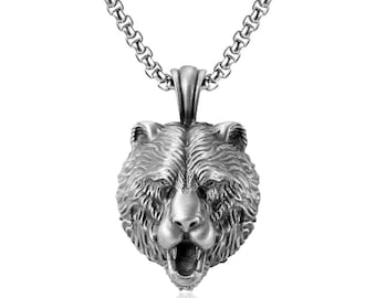 May include: A silver-coloured pendant necklace featuring a detailed bear head with an open mouth. The bear head is suspended from a chain with a textured link design. The pendant has a three-dimensional appearance against a white background.