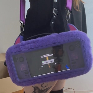 May include: A purple faux fur carrying case for a handheld gaming console. The case features a clear front window displaying a screen with the text 'Team Byte: The Backrooms'. It includes a purple adjustable shoulder strap with metal hardware clips.