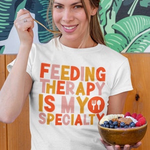 May include: A white t-shirt with the text "FEEDING THERAPY IS MY SPECIALTY" in orange, pink, and yellow letters. A fork and knife icon is next to the word "MY". The person is holding a bowl of fruit and granola.