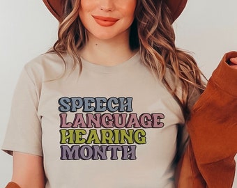 Può includere: Una donna indossa una maglietta beige con il testo "SPEECH LANGUAGE HEARING MONTH" in lettere retrò colorate. Indossa un cappello marrone e una giacca marrone.