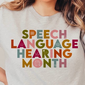 Puede incluir: Una camiseta gris claro con el texto "SPEECH LANGUAGE HEARING MONTH" en letras de bloque coloridas. La camiseta presenta varios iconos relacionados con el habla y la audición. La persona que lleva la camiseta está sonriendo.