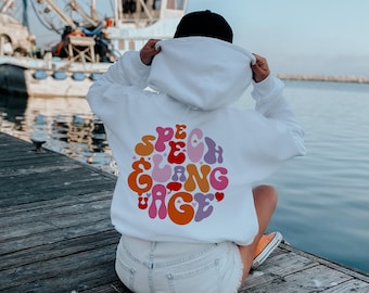 May include: White hooded sweatshirt with a colorful graphic on the back that says "Speech & Language". The person is wearing the sweatshirt and is sitting on a wooden dock by the water.