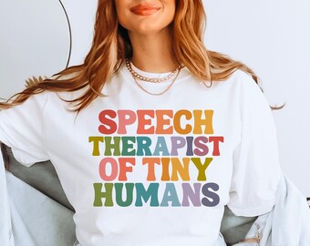 Op de afbeelding: Een wit sweatshirt met de tekst "SPEECH THERAPIST OF TINY HUMANS" in regenboogkleuren. De tekst is in een retro-lettertype stijl.