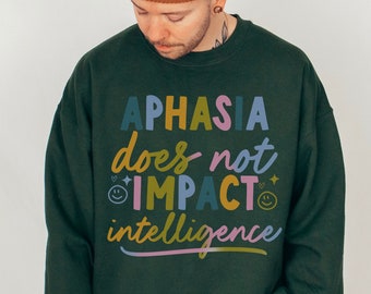 Puede incluir: Una sudadera verde oscuro con el texto "Aphasia does not impact intelligence" en letras de colores. El texto está rodeado de pequeños corazones y caritas sonrientes.