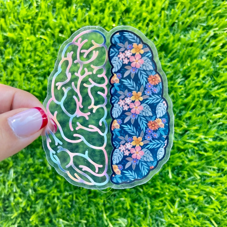 May include: A clear sticker in the shape of a brain, split into two halves. One side has a holographic, brain-like pattern. The other side features a floral design with blue, pink, and yellow flowers on a dark blue background. The sticker is held against a green background.