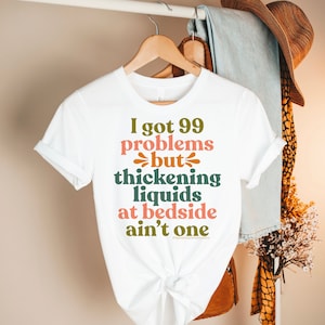 Puede incluir: Camiseta blanca con un dicho divertido en texto verde, naranja y rosa. El texto dice "Tengo 99 problemas, pero los líquidos espesantes en la cama no son uno".