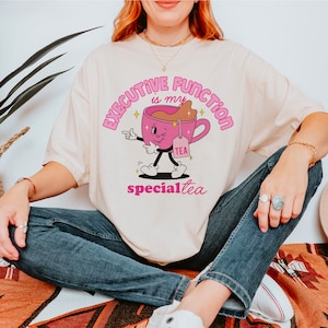 Puede incluir: Camiseta color crema con un personaje de taza de té de dibujos animados y el texto "EXECUTIVE FUNCTION is my TEA specialtea" en rosa. La taza de té es rosa con líquido marrón y una bolsita de té. La camiseta la lleva una persona.
