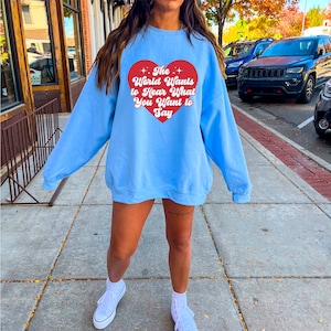 Puede incluir: Una sudadera azul claro con un gráfico de corazón rojo que dice "The World Wants to Hear What You Want to Say".