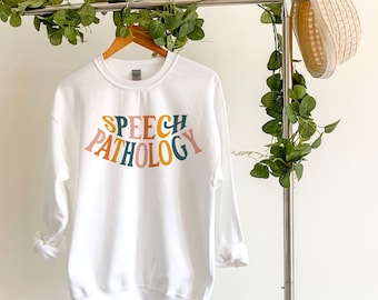 May include: White sweatshirt with the text "SPEECH PATHOLOGY" in a rainbow arch design. The sweatshirt is hanging on a clothing rack.