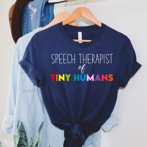 Puede incluir: Una camiseta azul marino con el texto "Speech Therapist of Tiny Humans" en blanco, con la palabra "Tiny" en colores arcoíris.