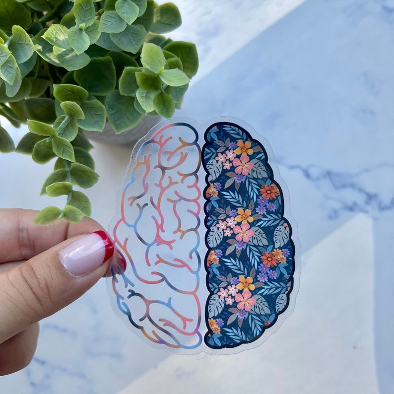 May include: A clear sticker in the shape of a brain, with one side featuring a colorful brain pattern and the other side adorned with a floral design in shades of orange, pink, and blue. The sticker is held by a hand.