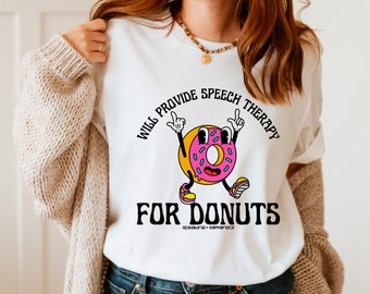 Può includere: Maglietta bianca con un motivo a ciambella rosa e blu. La ciambella indossa le scarpe e ha le braccia alzate in aria. Il testo sulla maglietta recita "Will provide speech therapy for donuts speaking = samantics".
