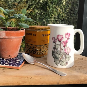 Puede incluir: Una jarra de cerámica blanca con un diseño floral de flores de ciclamen rosas y hojas verdes. La jarra tiene un asa y está sentada sobre una superficie de madera. En el fondo hay una pequeña maceta de terracota con una planta suculenta verde, junto con una cuchara de plata y una lata de té amarilla que dice "Jasmine Tea" y "Thé au Jasmin".