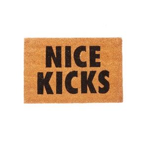 Pode incluir: Um tapete de coco castanho com o texto "NICE KICKS" em letras pretas.