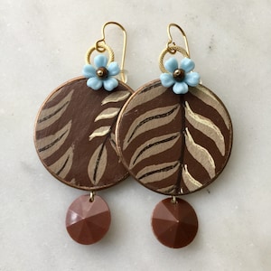 Puede incluir: Un par de pendientes de color marrón y beige con estampado de hojas, un dije de flor azul claro y una cuenta facetada marrón.