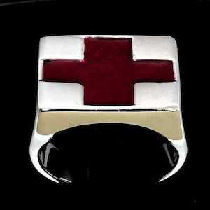 May include: A silver ring with a rectangular face featuring a red cross design. The cross is centered on a white background, with a gold-coloured band at the base of the face. The ring has a polished, reflective finish.